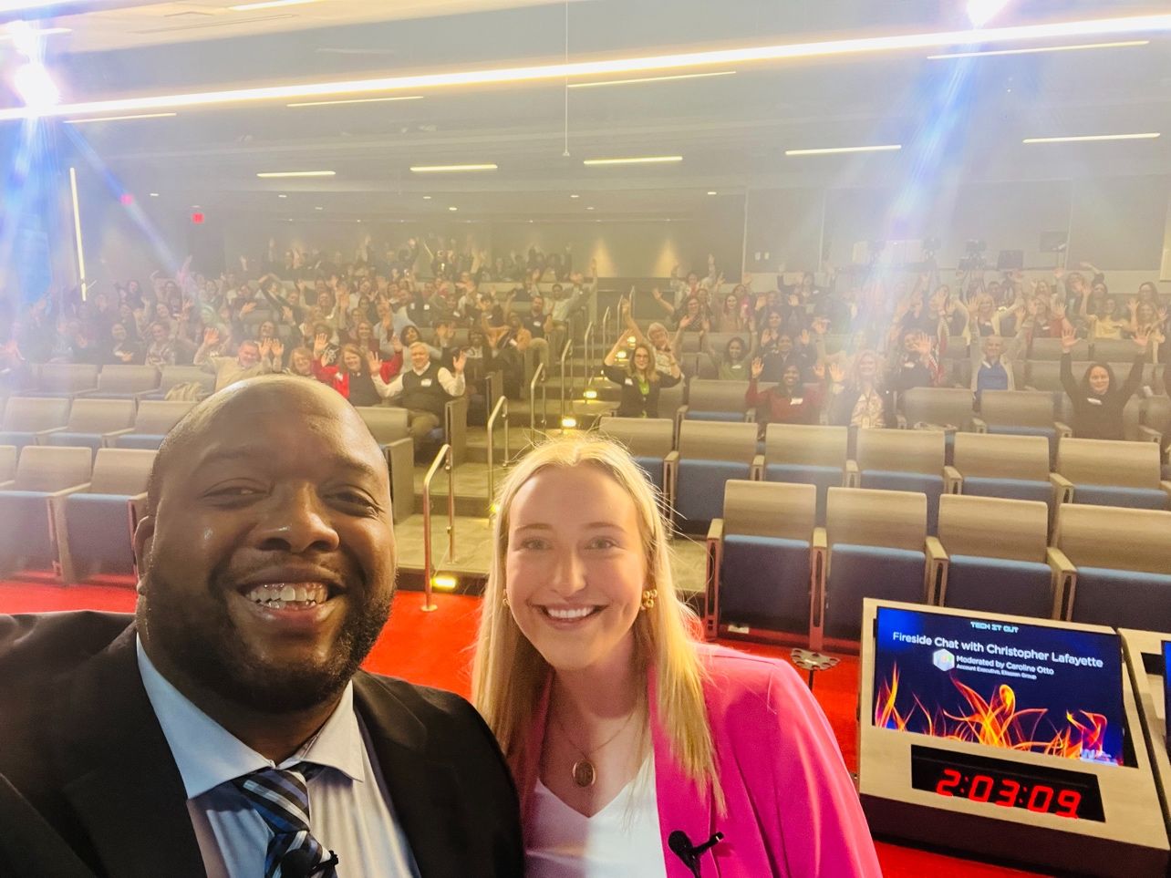 Two people in a selfie at the front of an auditorium, with the audience waving and a screen reading “Tech It Out” and “Fireside Chat with Christopher Lafayette.”