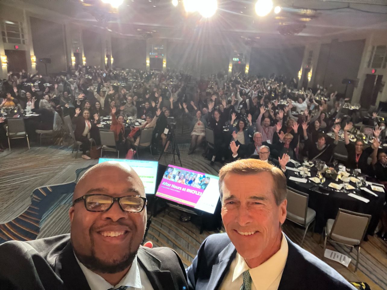 Two people in a selfie at the front of a conference ballroom, with the audience waving behind them.