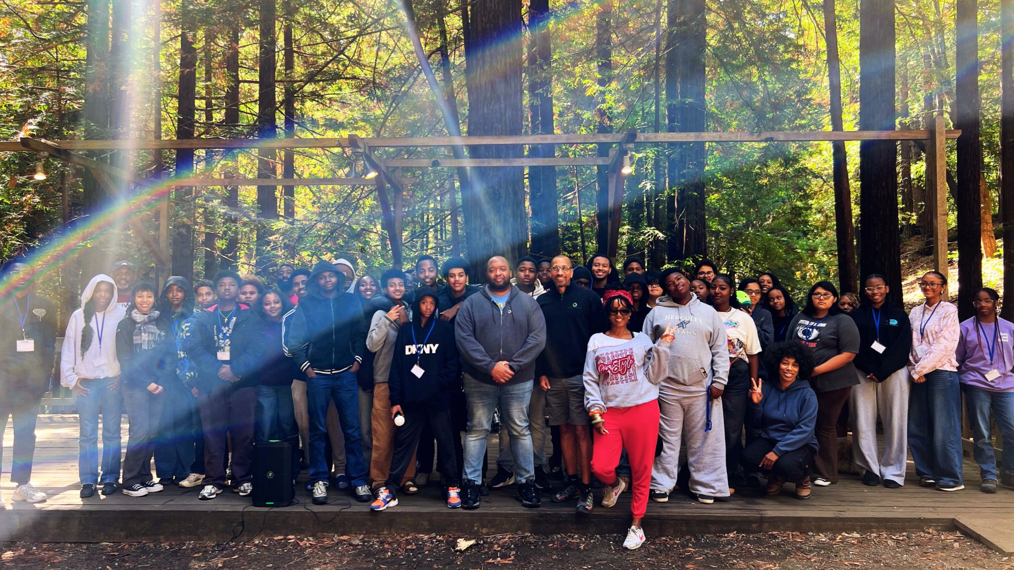 A group photograph on a wooden platform surrounded by tall trees and sunlight.