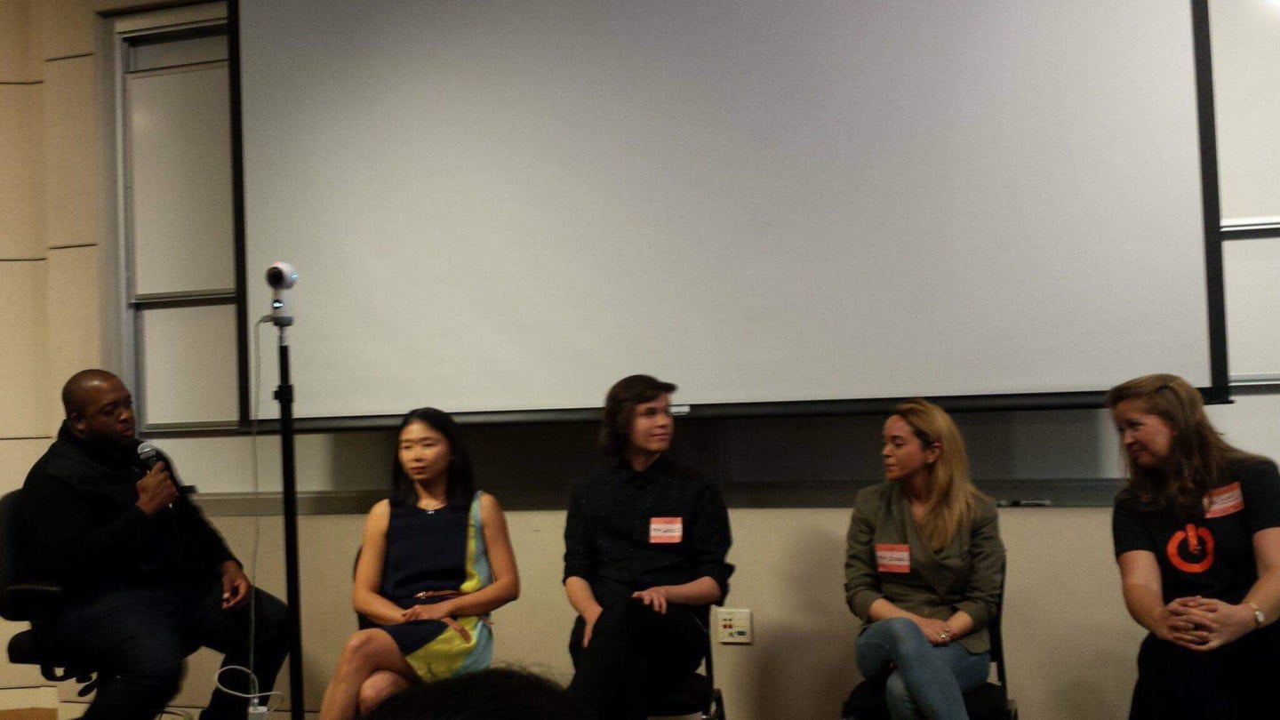 Christopher Lafayette holds a microphone at the left of a five-person panel beneath a large projection screen at Stanford.