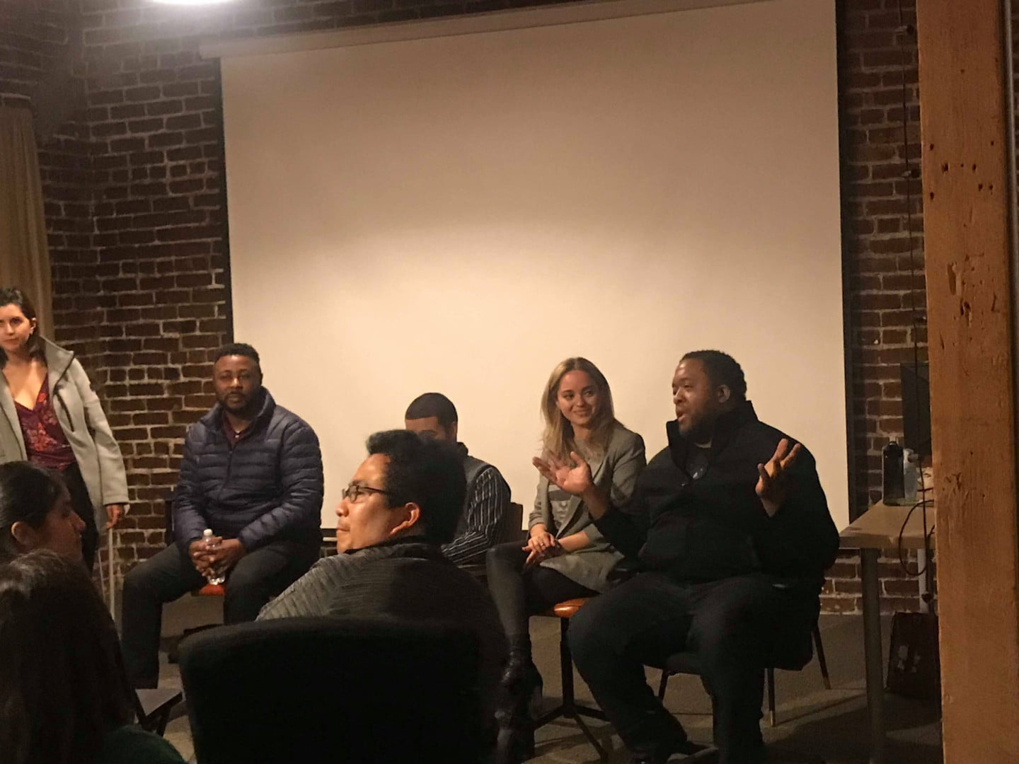 Christopher Lafayette gestures while seated with fellow panelists in a brick-walled room, with audience members in the foreground.